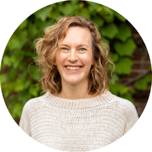 woman with cream sweater standing in front of greenery. She has a smile on her face.